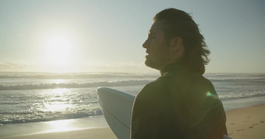 A man standing on the beach holding a surf board looks off in to the distance