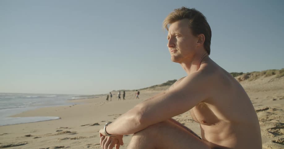A shirtless man sitting on the beach looking at the ocean