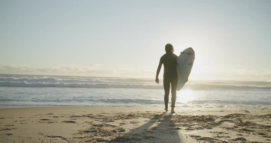 Surfer wearing a black wet suit on a beach carries a surfboard to the ocean at sunset