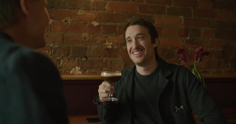 Two men sitting while smiling and talking as one man holds a drink