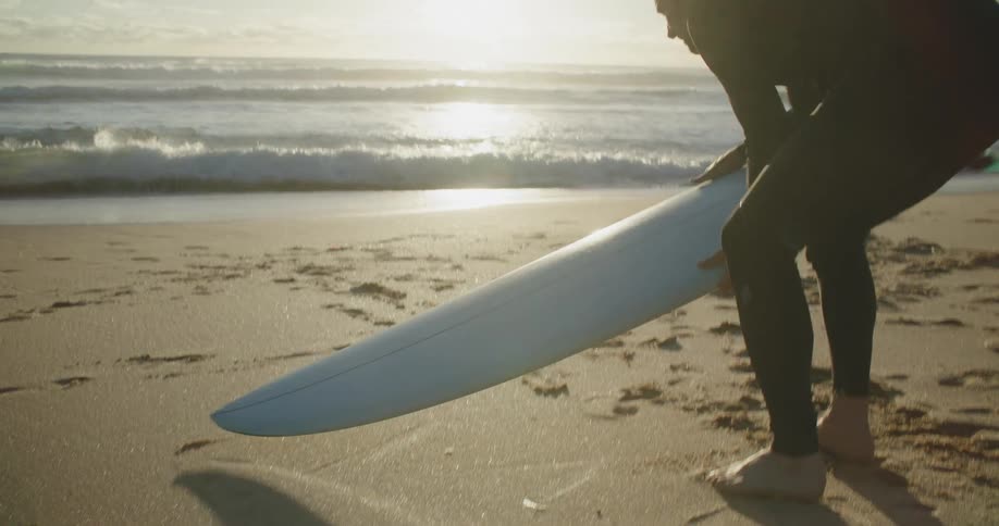 Man wearing a black wet suit picks up a surfboard from the sand of a beach near the ocean at sunset