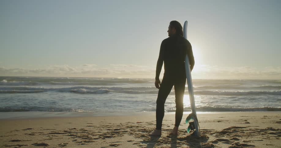 A man holding a surfboard stands contemplatively on a beach on a sunny day