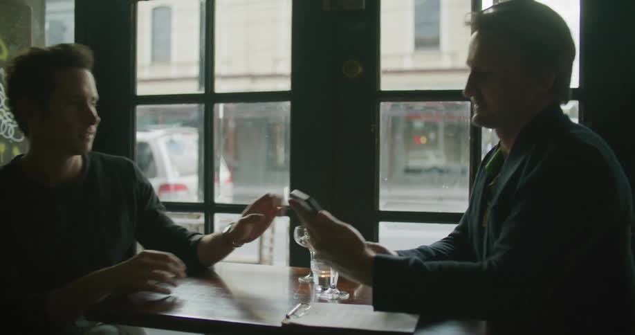 Two men are sitting at a table in a bar drinking and looking at a cell phone