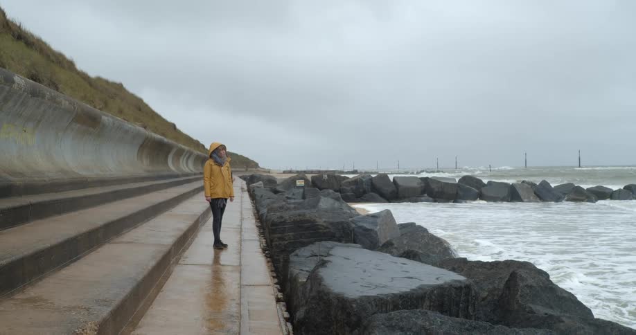 A woman standing on a concrete slab watches wave crash in to rocks on the shoreline