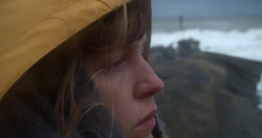 Young blonde woman wearing a yellow windbreaker stands on the rocks next to the ocean