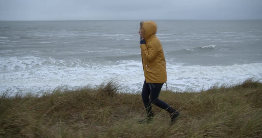 A woman holds her raincoat hood closed as she walks through tall grass along a cliff overlooking the ocean