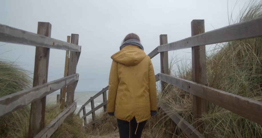 Woman wearing a yellow raincoat walks on a fenced road close to the ocean