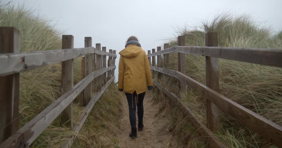 A woman in a yellow parka walks on a dirt trail that runs between two fenced off fields near the ocean