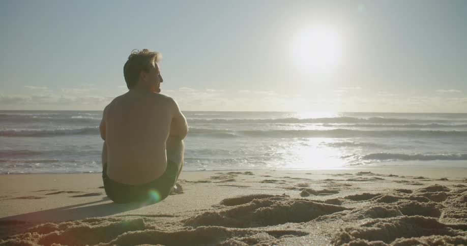 Man wearing swimming trunks sits on the sandy beach near the ocean on a sunny day