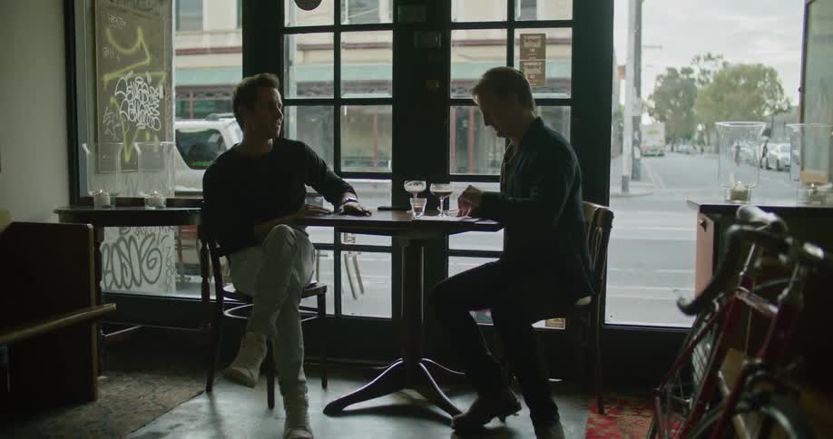 Two men sit at a table in a bar and have a drink and talk and look at papers as people walk past outside