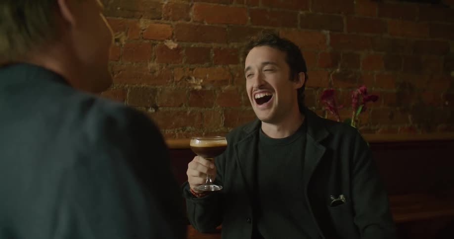 A smiling man holding a full drinking glass talks with another man indoors