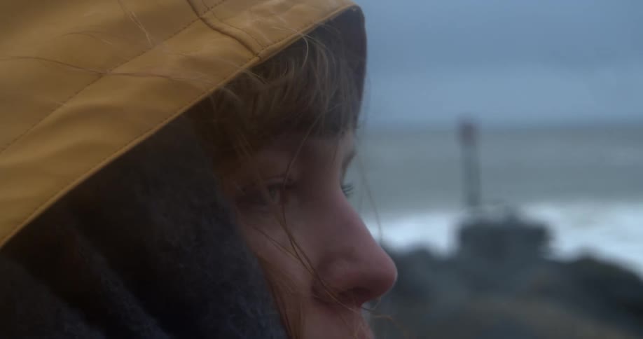 A woman wearing a yellow raincoat stands outside next to an ocean at daytime and turns to look behind her