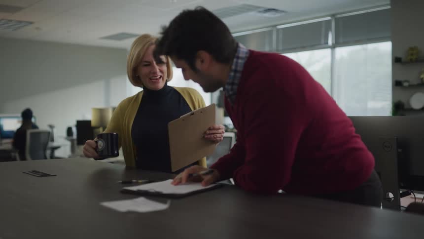 A man leans over a document that he is marking when a female walks over to review it with him