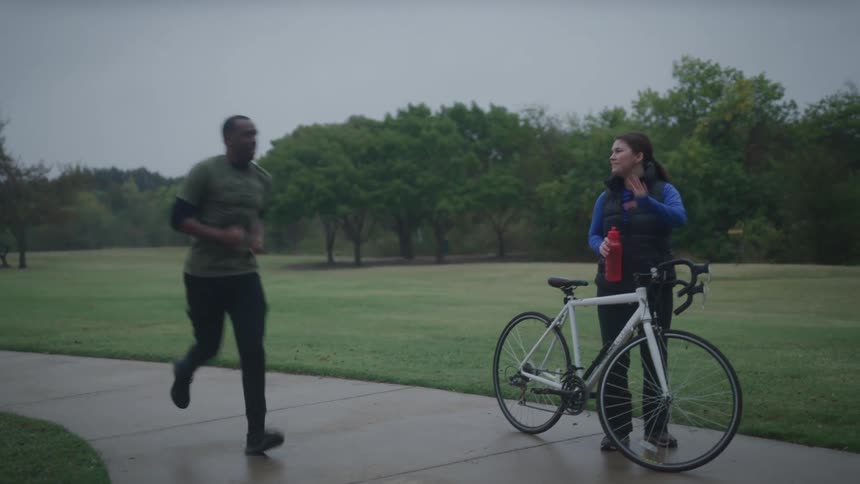 A woman holds her bike and takes a drink of water and waves at a man as he jogs by