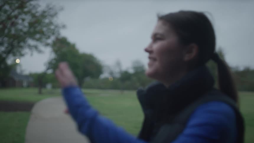 A woman drinking a from a water bottle while sitting outdoors on a cloudy evening waves at a man as he jogs by