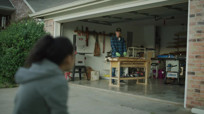A grandfather age veteran doing carpentry work in his garage stops and waves at a little girl in the front yard.