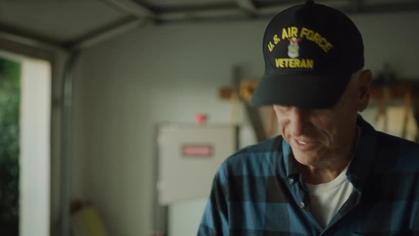 A grandfather age veteran doing carpentry work in his garage stops and waves at someone in the front yard.