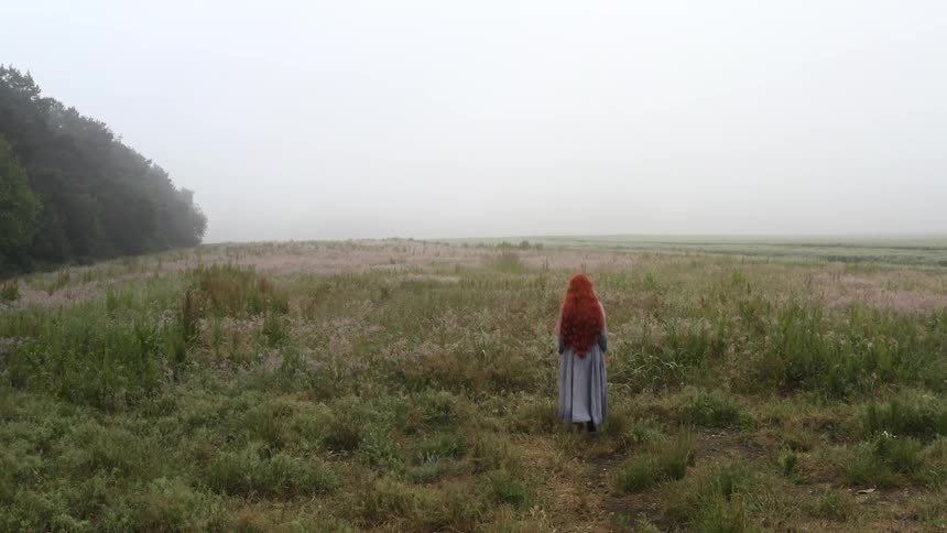 Woman with long red hair wearing a dress walking through field