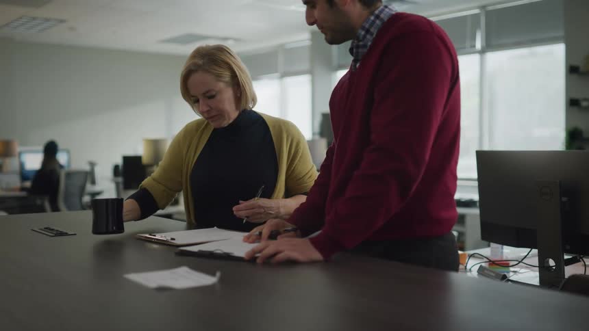 Woman in yellow sweater drinks coffee while talking to a coworker in the office