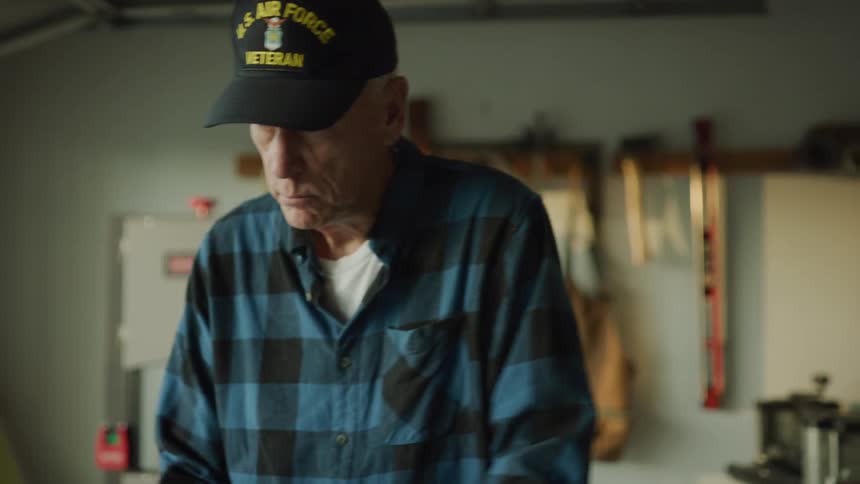 A grandfather age veteran doing carpentry work in his garage stops and waves at someone in the front yard.