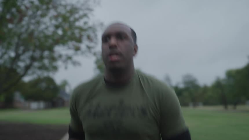 A man jogging on a path in a park by a neighborhood waves at a woman by her bike