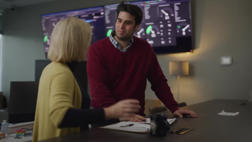 Woman holding a mug walks over to a man standing at a desk and puts down her coffee and talks to him in an office at daytime