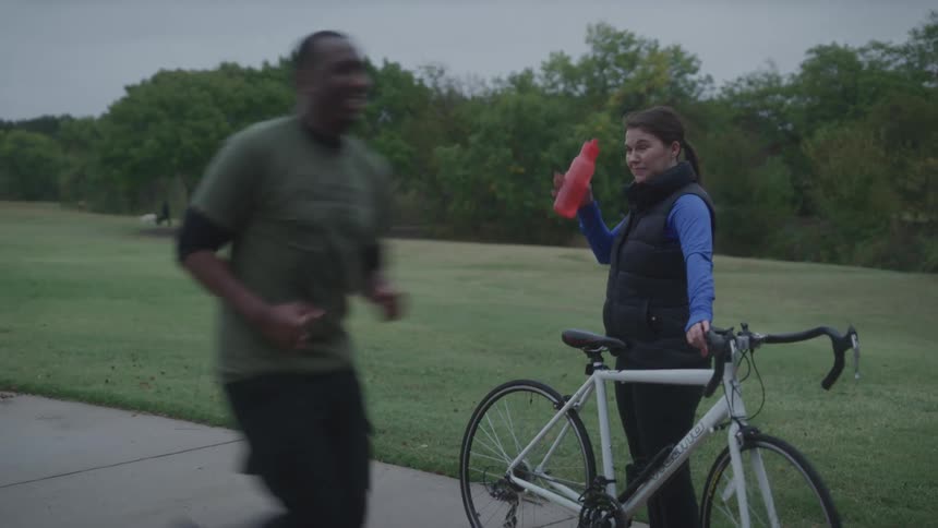 A woman stands by her bike drinking from a water bottle in a park as a man jogs past