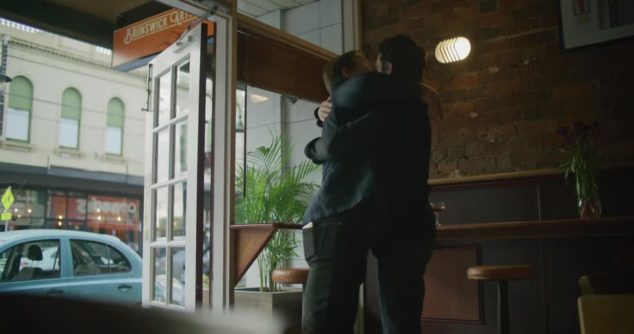 A man sitting at a restaurant table having a drink stands up when his friend walks in and they hug