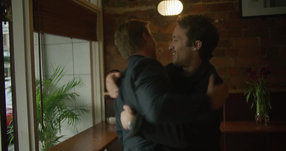 A man rises from his seat to greet another man with a hug before inviting the other man to take a seat indoors during the day