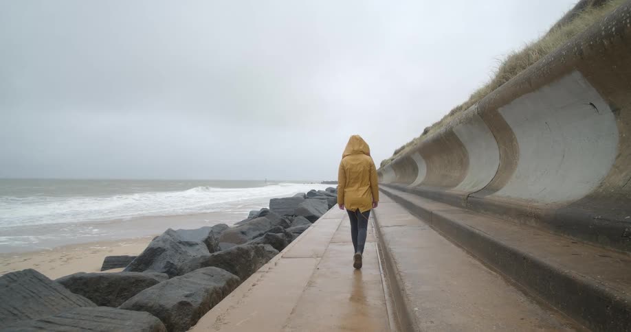 A person walks alone on a sidewalk next to the ocean wearing a raincoat