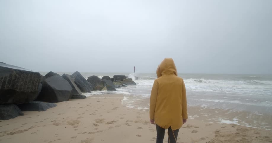A woman wearing a hooded coat walks across a sandy beach towards a line of large stones on a cloudy day