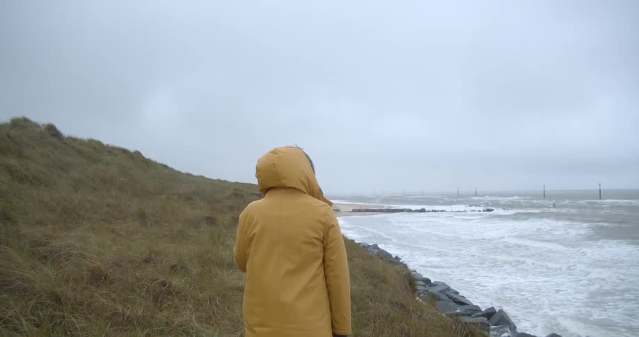Person wearing yellow hooded jacket walks along coastal bluff as the waves roll in