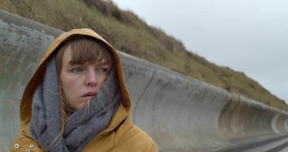 A woman wears a gray scarf and yellow raincoat over her head as she walks along a retaining wall below a hill