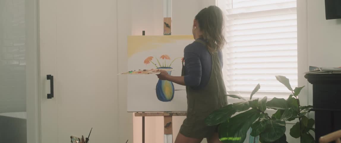 A young woman carrying a palette and paint brush approaches a canvas on an easel and continues a painting
