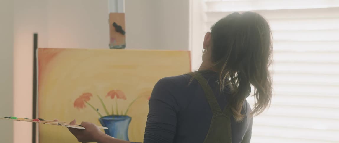 A woman stands at an easel painting on a canvas indoors