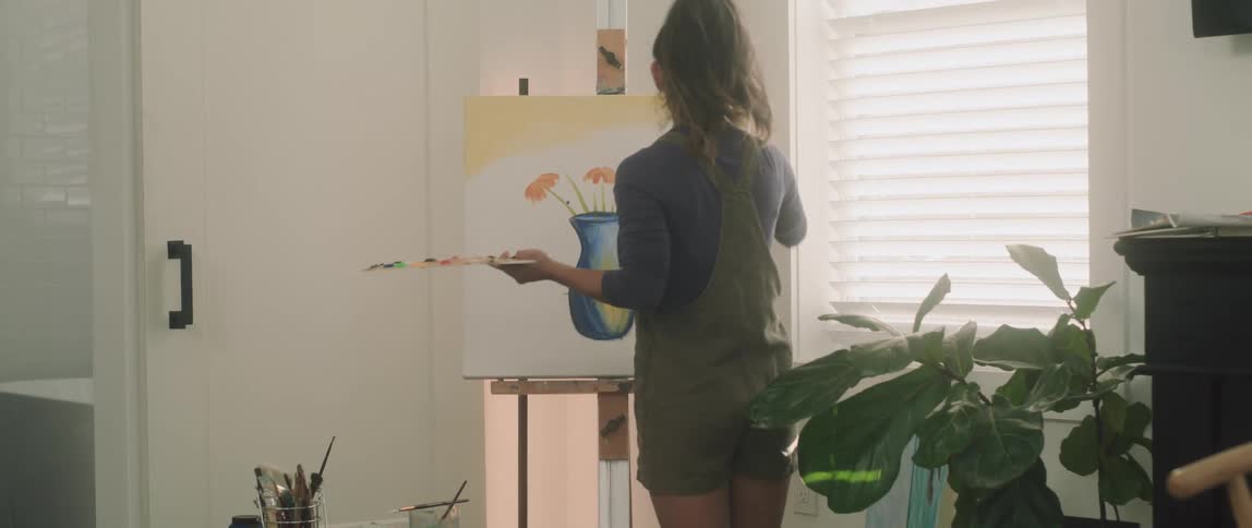 A young woman paints a drawing of a vase on an easel set up next to the window in her apartment