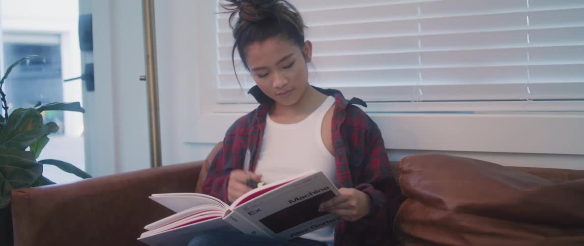 A young woman sits down on her couch with a textbook and pen in hand to study