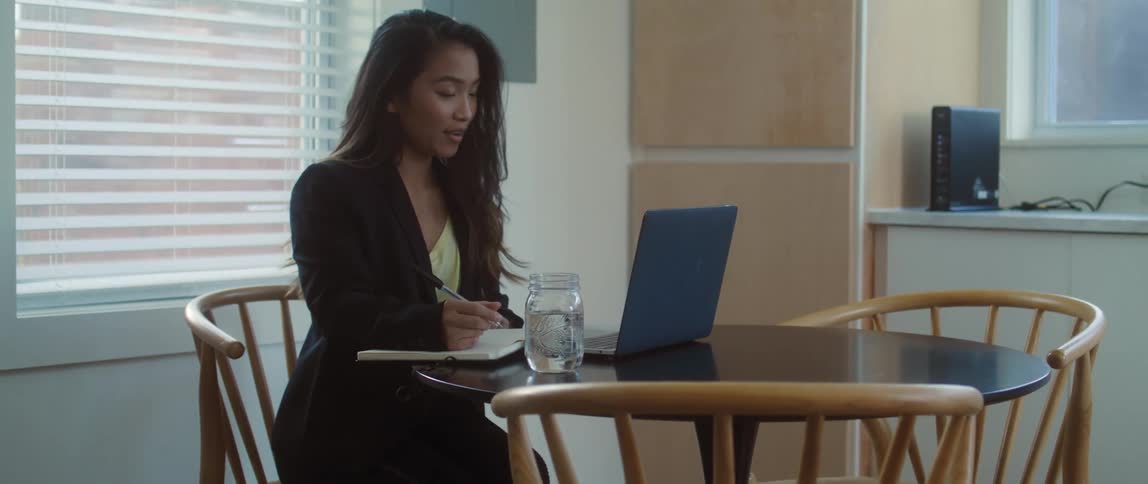 A woman sits at a dining room table at home taking notes with a notebook on a laptop video call during the day