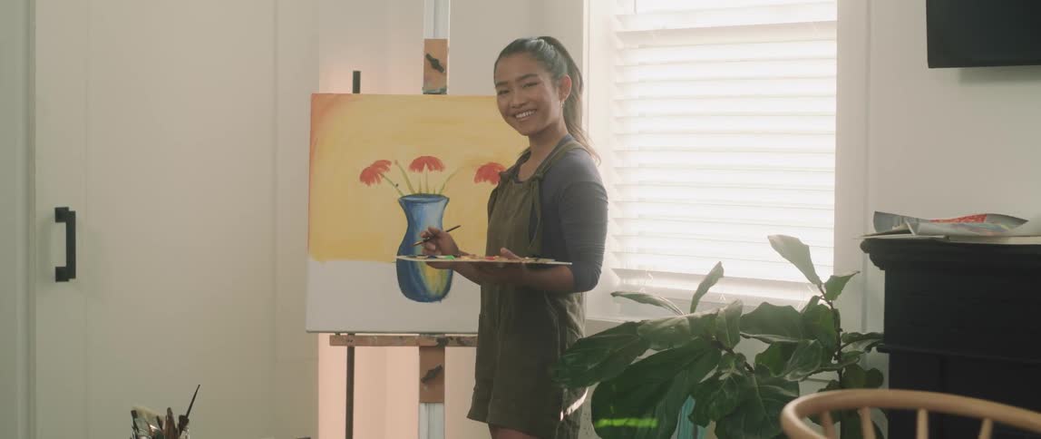 A woman in a room holding a paint palette and a paint brush with a painting in the background