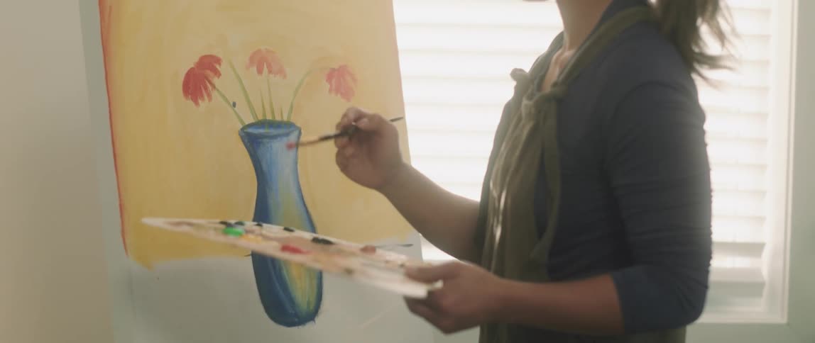 A woman is painting flowers in a vase on a canvas