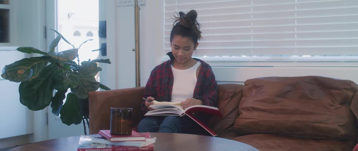 A teenage girl at home walks to a couch and sits down and opens a book to read