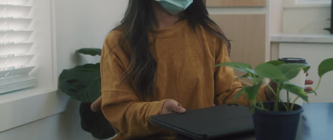 Woman wearing a face mask closes a laptop and stands up from a table
