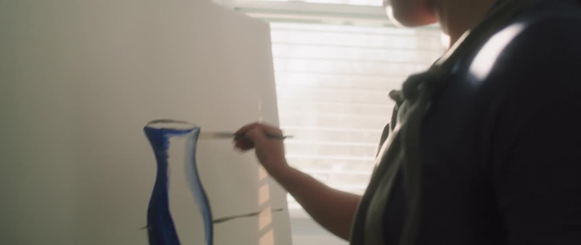 An asian teenage girl stands at an easel painting a blue vase on a canvas