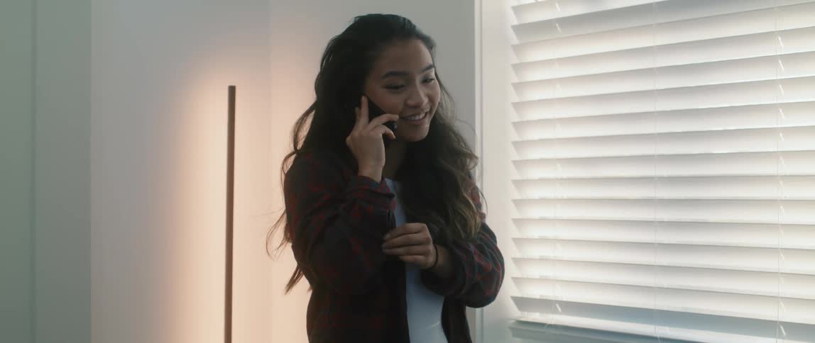 A young asian woman smiles while speaking on her smartphone while standing near a window in a building during the day