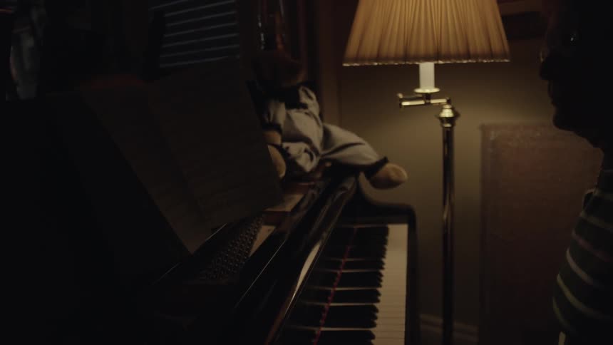 An older woman sits down in front of a piano lit with a single lamp