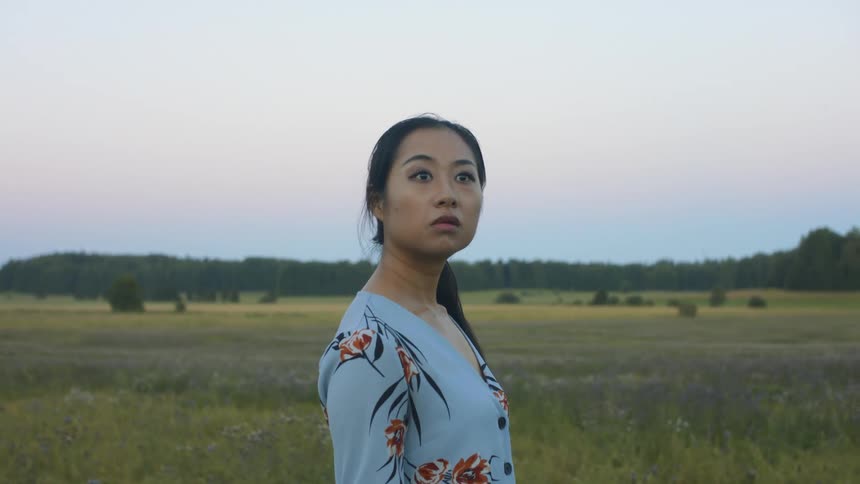 A young asian woman with her back towards the camera in a grassland suddenly turns to look at something out of frame with a startled expression on her face