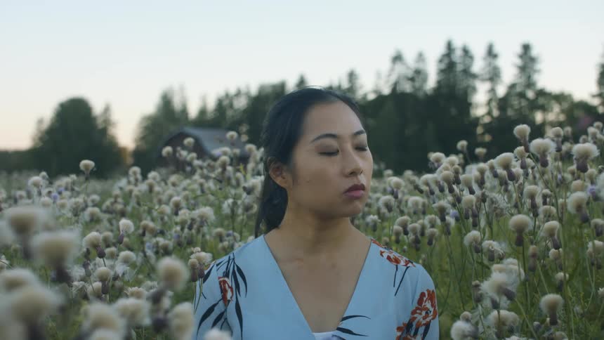 A woman standing in a grassy field with flowers with a building trees the sky and clouds in the background