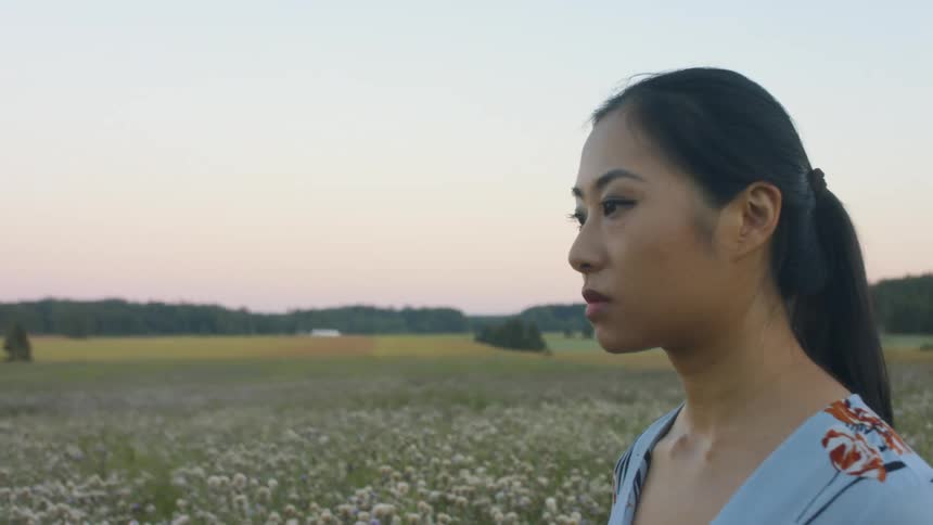 A young asian woman walks through a grassland with a solemn expression on her face in the evening