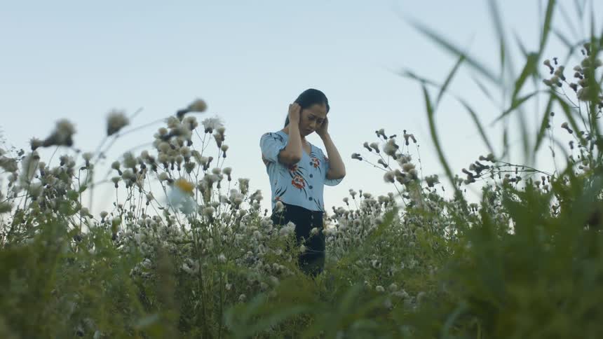 A woman stands in a field outside and places her hands at her ears as she looks around