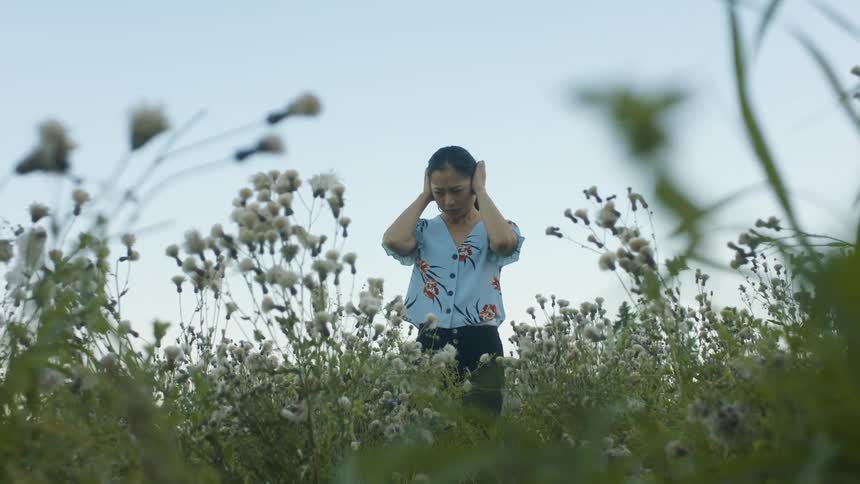 A woman standing in a grassy field covering her ears with the sky and clouds in the background
